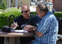 Two men of color with glasses standing at a table outside writing oin a notebook. 