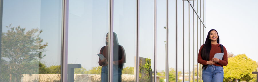 Female student walking outside next to a reflective building, with a reflection of the student in the window