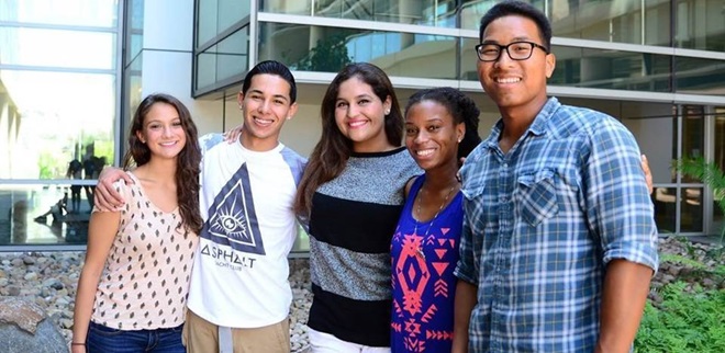 five smiling students in front of a glass building