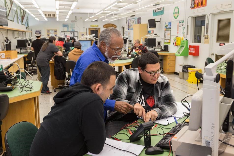 Male appearing professor helping twomale appearing students with work at a computer. Four other students appear in the background distance