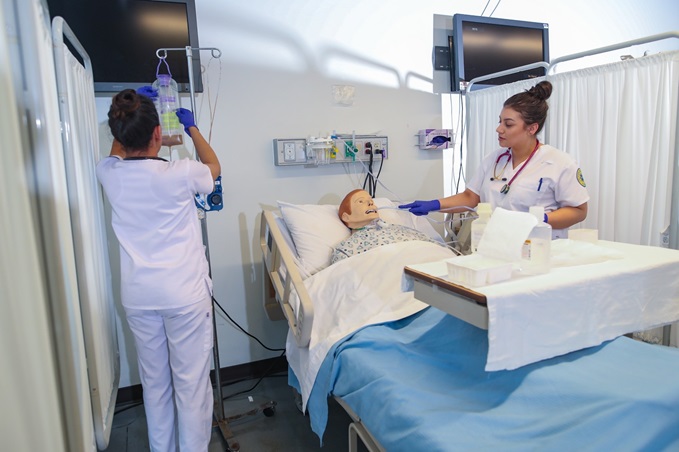 Hospital room with patient dummy in bed, one worker standing by the bed and one worker hanging an IV drip to a cart