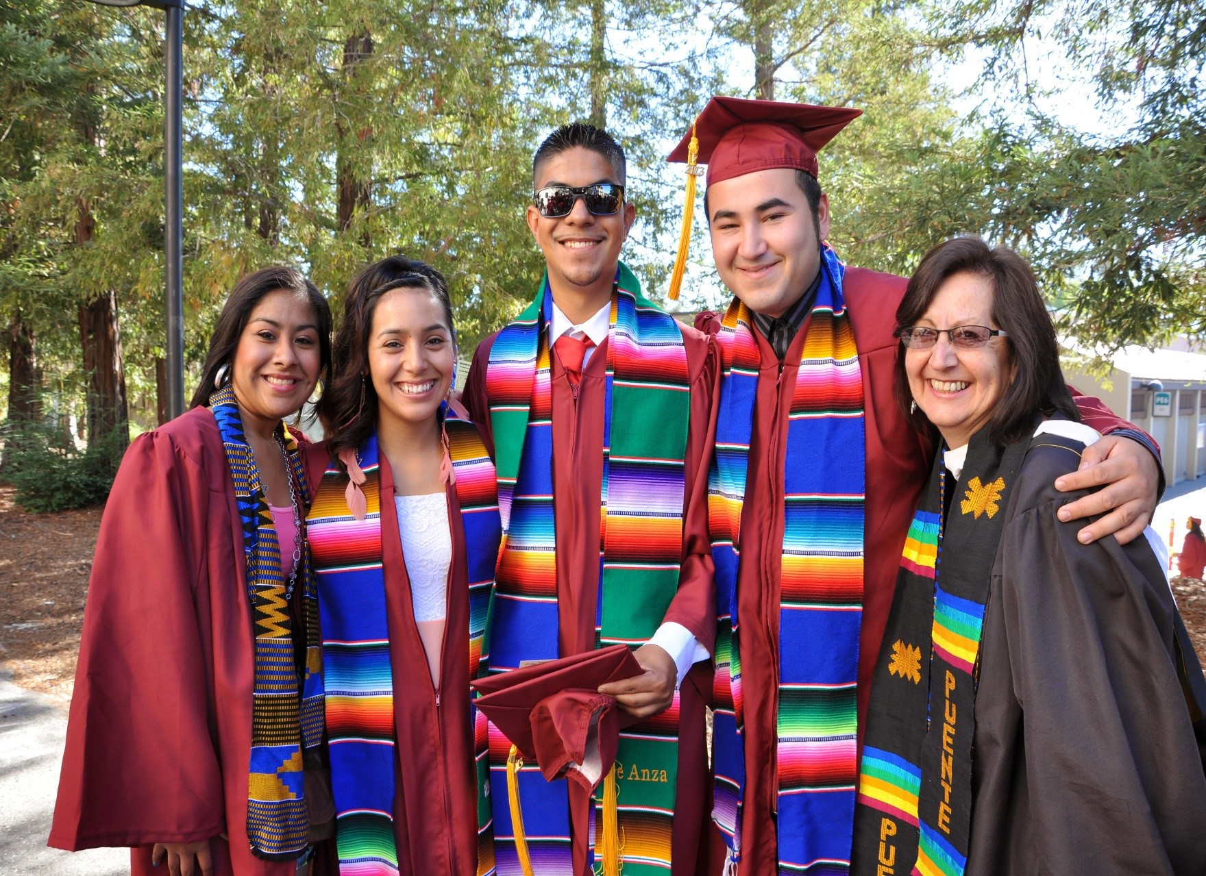 Five graduating students: four in red gowns and one in black with colorful sashes