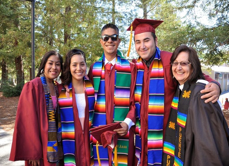 Five graduating students: four in red gowns and one in black with colorful sashes