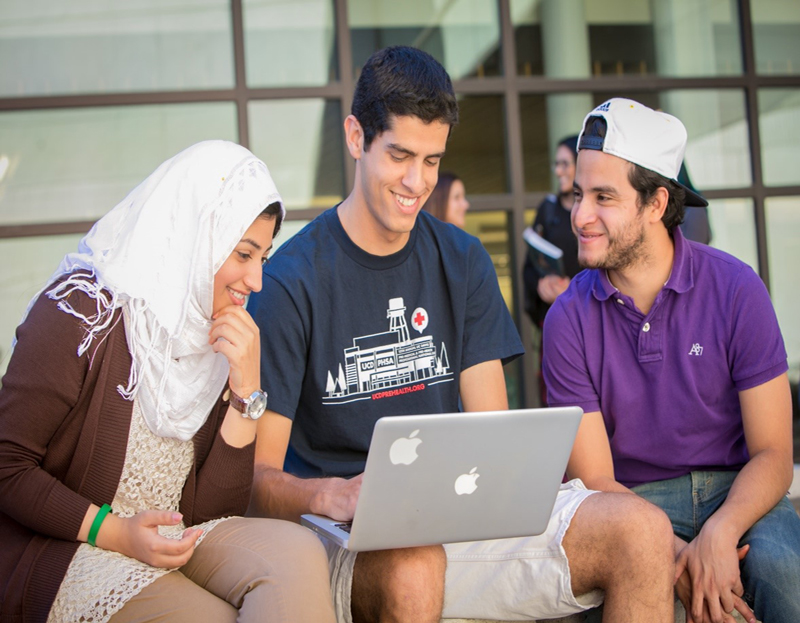 Three students sitting outside looking at a Mac laptop. Student at far lap appears female with a hijab. middle student appears male, student at far right appears male with a white baseball cap.
