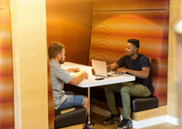 Two male-appearing people at a booth. One has a laptop on the table in front.