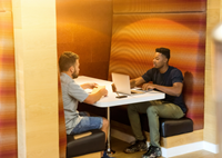 Two male-appearing people at a booth. One has a laptop on the table in front. 