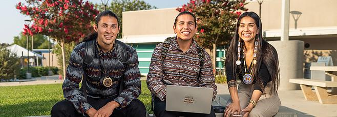 Three smiling native american students sitting outside on a community college campus. The middle student has a laptop on their lap.
