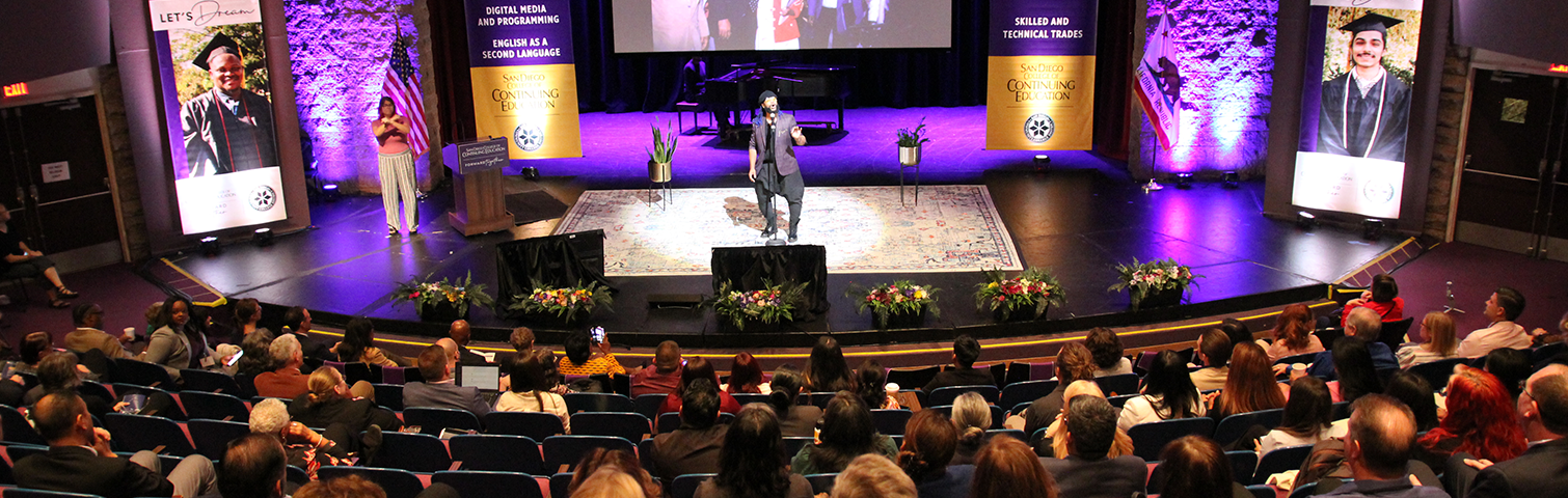 Stage with a person speaking on it surrounded by curved rows of chairs filled with people