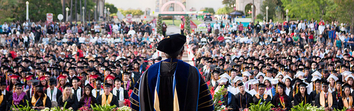 a photo taken from the stage with rows of audience in front, and the back view of a person speaking and wearing a graduation cap and gown.