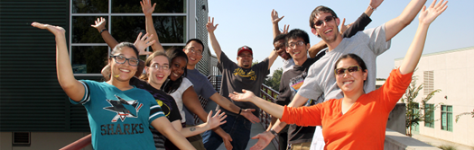 Photo taken outside in the front of buildings with nine students facing the camera with their hands in the air.